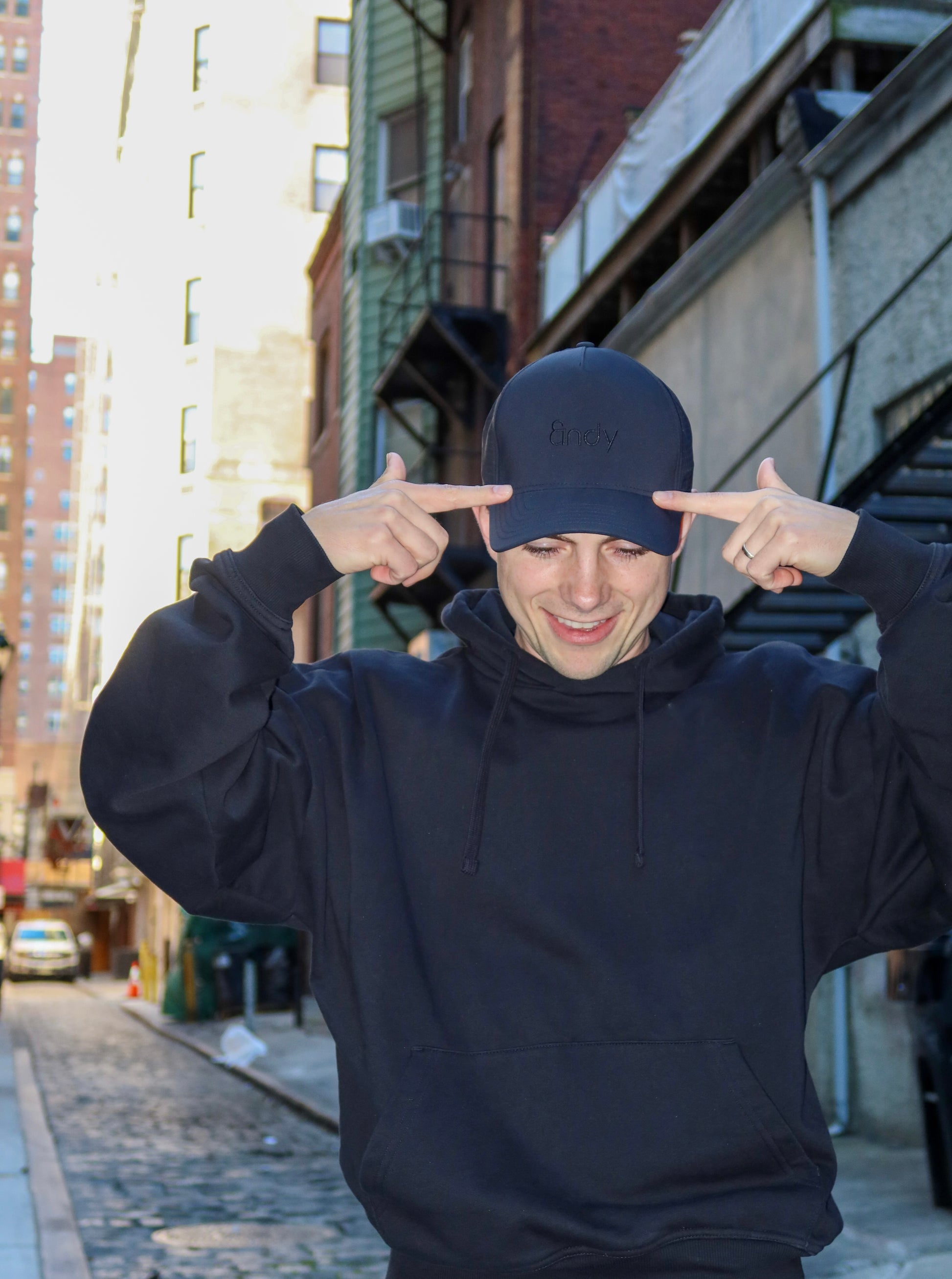 Person wearing a black hoodie and black cap pointing to their head on a city street.