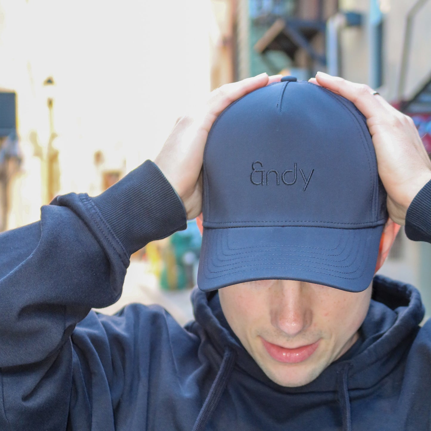 Person wearing a black cap with 'Andy' branding, holding it above their head.