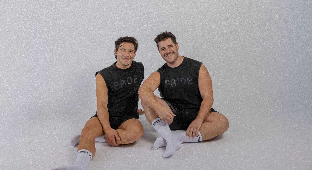 Two men wearing black sleeveless shirts with 'PRIDE' text, sitting on a light gray background.